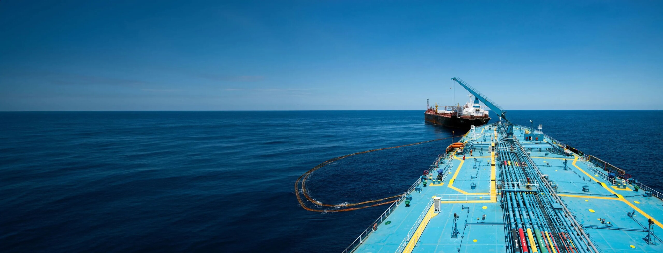 View from the deck of a large cargo ship with blue-painted surface and pipelines, looking toward another vessel ahead on calm open sea under a clear sky.